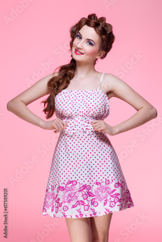 Beautiful young woman with long red braided hair and bright colourful makeup wearing dress standing on pink background. Pin up style. Copy space.