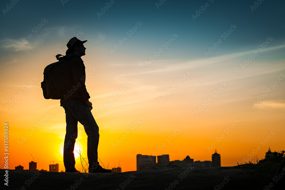 Obraz premium Traveler silhouette watching amazing sunset. Young casual man with backpack and cowboy hat standing alone on hill above evening cityscape. Lifestyle Travel Concept Outdoor Background. Kiev, Ukraine