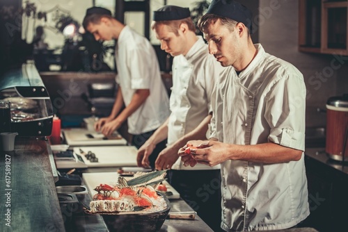 Food concept. Three young chefs in white uniform decorate ready dish in restaurant. They are working on maki rolls. Preparing traditional japanese sushi set in interior of modern professional kitchen photo