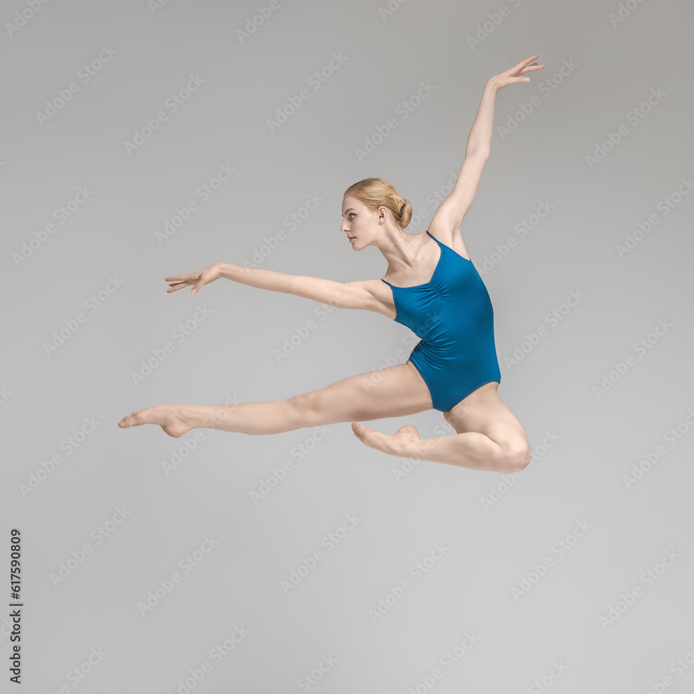 Delightful ballerina posing in the jump on the gray background in the studio. She wears a blue leotard. Her legs and right arm stretched forward, left arm stretched upward. Shoot from the side.