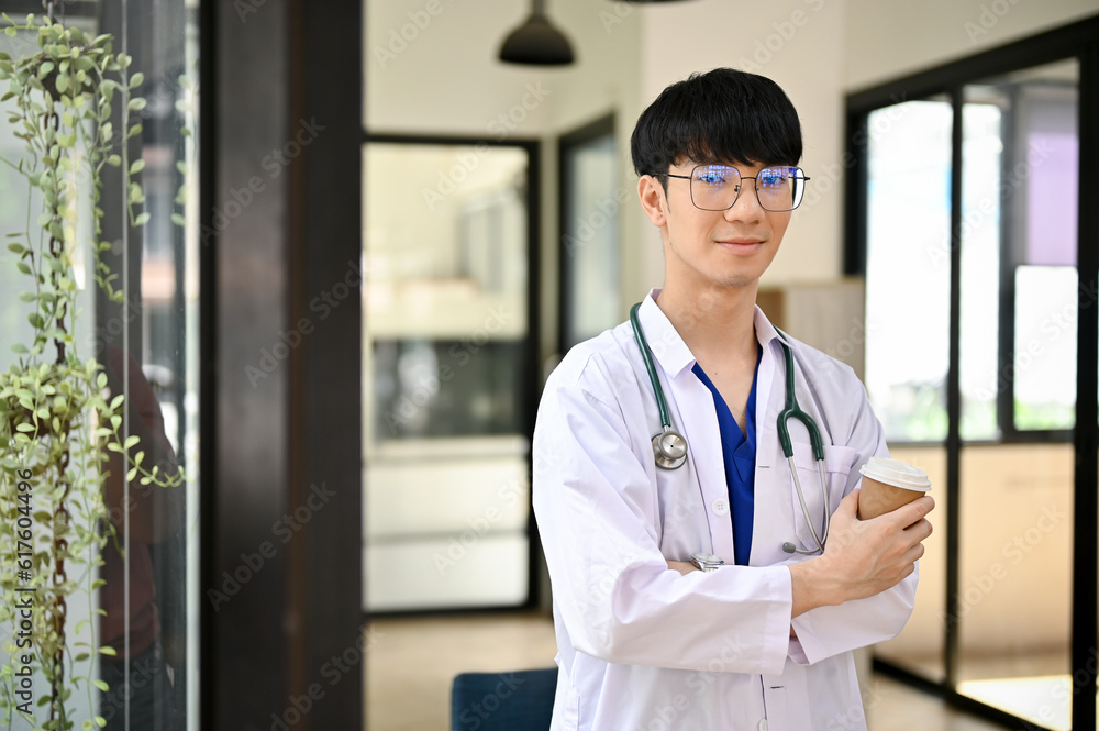A male doctor in a uniform with a stethoscope stands in the hospital corridor with a coffee cup A male doctor in a uniform with a stethoscope stands in the hospital corridor with a coffee cup