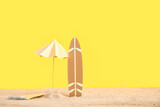 Creative composition with mini surfboard, umbrella and blanket on sand against yellow background