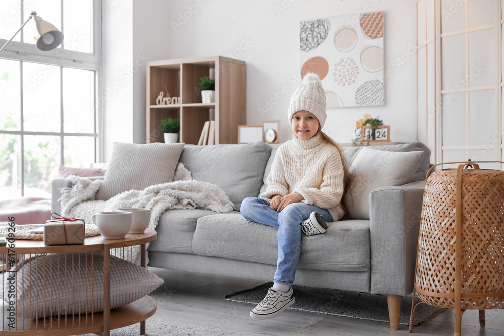 Cute little girl in knitted hat and sweater sitting on sofa at home Cute little girl in knitted hat and sweater sitting on sofa at home