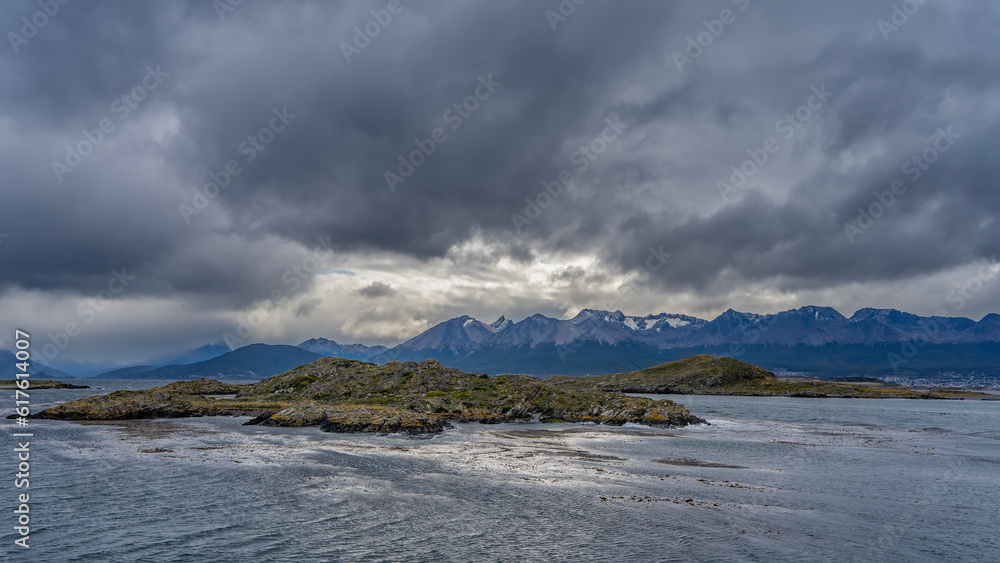 A rocky island with sparse vegetation in the sea strait. A picturesque snow-capped mountain range against a cloudy sky. Argentina. Tierra del Fuego Archipelago. Beagle Channel