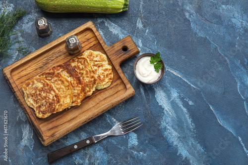 Wooden board of tasty zucchini fritters with sour cream on blue background