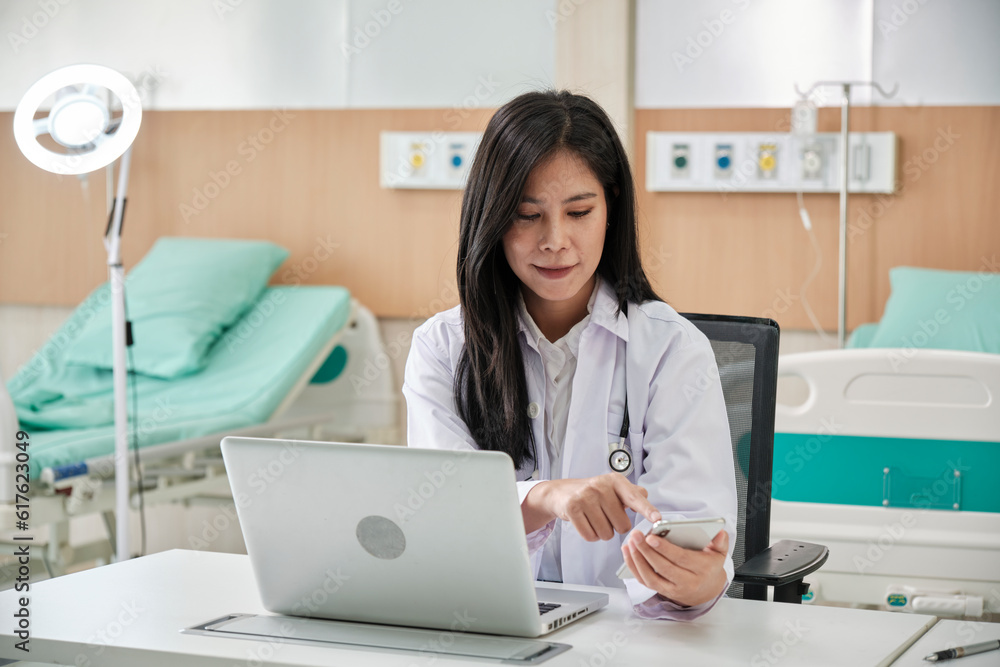 Young beautiful Asian female doctor online working with laptop and smartphone on medical consultation application via internet at working desk in hospital clinic. Young beautiful Asian female doctor online working with laptop and smartphone on medical consultation application via internet at working desk in hospital clinic.