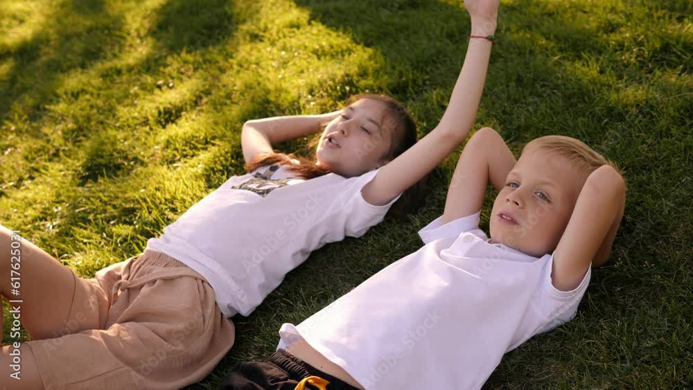 Brother and sister lie in the summer in the park on the grass with their hands behind their heads. Children rest on the green grass in the city park. Brother and sister lie in the summer in the park on the grass with their hands behind their heads. Children rest on the green grass in the city park.
