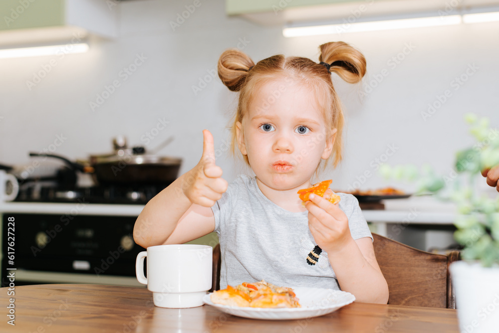 Sad little caucasian girl eating fast food while sitting at the table in the kitchen and showing like gesture Sad little caucasian girl eating fast food while sitting at the table in the kitchen and showing like gesture