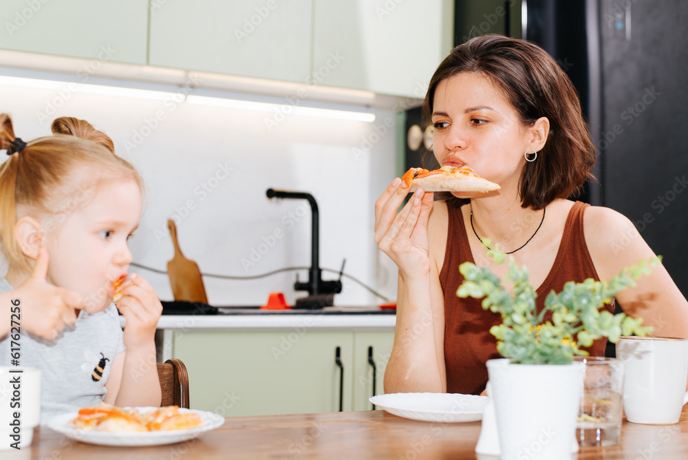 Mom and daughter eat together in kitchen at home, family lunch. Mother looking at her baby girl sitting at table Mom and daughter eat together in kitchen at home, family lunch. Mother looking at her baby girl sitting at table