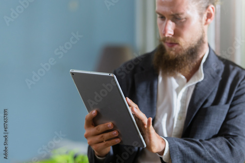 Close up selective focus at tablet in hand of businessman. Handsome businessman with beautiful bearded standing working online using tablet at window in his office. browsing information on internet.