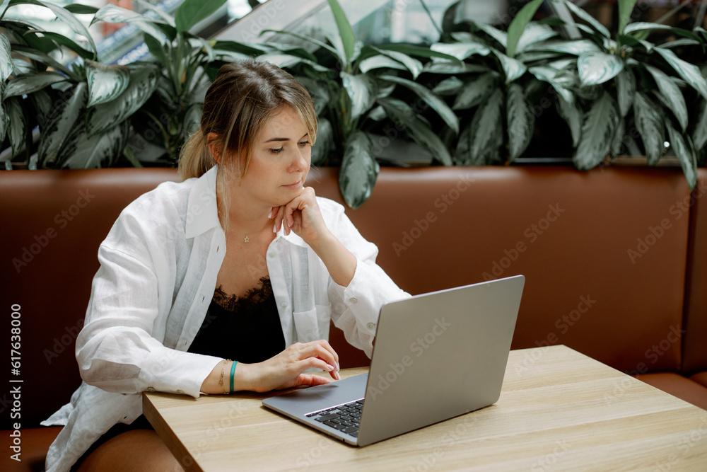 Concentrated lady freelancer lean head on hand working with laptop sitting on couch at cafe table surrounded by plants. Concentrated lady freelancer lean head on hand working with laptop sitting on couch at cafe table surrounded by plants.