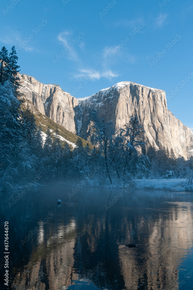 The rocky surface of the Sierra Nevada mountains, reflecting early morning sunlight, being reflected in Merced river, on an early winter, snow-covered landscape in Yosemite national park.