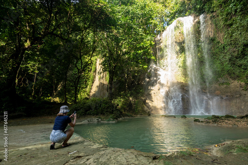 An asian tourist in a blue top and jorts taking a picture of a waterfall. At Kawasan falls in Balilihan, Bohol.
