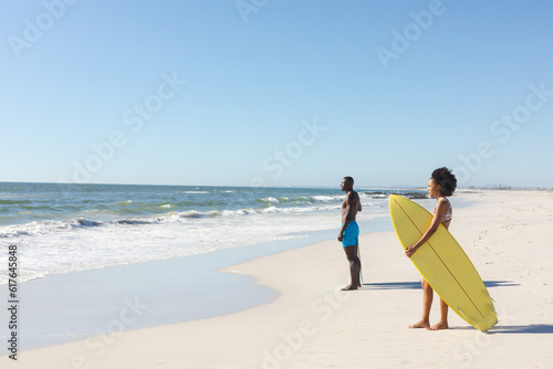 African american man and woman with surfboards standing on sunny beach looking to sea, copy space
