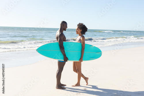 Happy african american couple holding surfboard smiling at each other standing on sunny beach