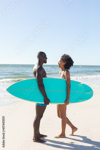 Happy african american couple holding surfboard smiling at each other on sunny beach, copy space