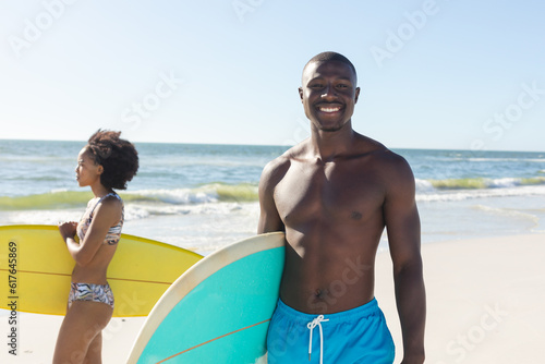 Portrait of happy african american man holding surfboard standing on sunny beach with female surfer