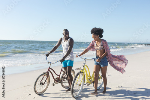 Happy african american couple walking with bikes on sunny beach by sea