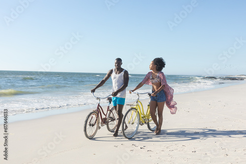 Happy african american couple walking with bikes on sunny beach by sea