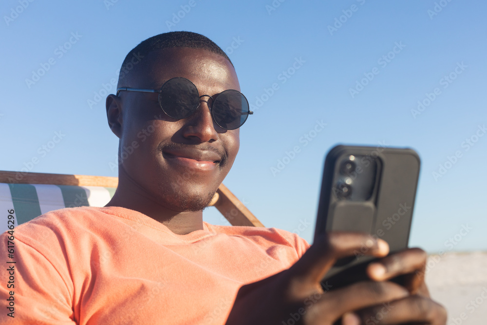 Happy african american man in sunglasses sitting in deckchair using smartphone on sunny beach Happy african american man in sunglasses sitting in deckchair using smartphone on sunny beach