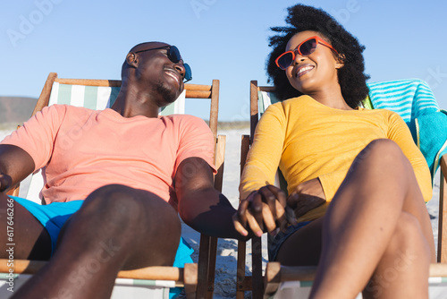 Fototapeta Happy african american couple sitting in deckchairs holding hands on sunny beach