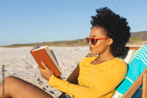 Happy african american woman in sunglasses sitting in deckchair reading book on sunny beach