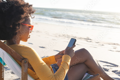 Happy african american woman in sunglasses sitting in deckchair using smartphone on sunny beach #617646289