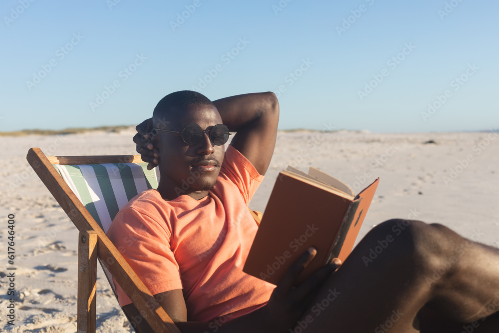 © wavebreak3 - Happy african american man in sunglasses sitting in deckchair reading book on sunny beach