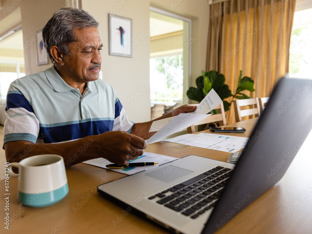 Senior biracial man doing paperwork and using laptop in dining room Senior biracial man doing paperwork and using laptop in dining room