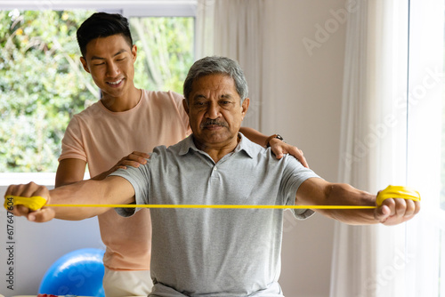 Happy diverse male physiotherapist advising and senior male patient using exercise band