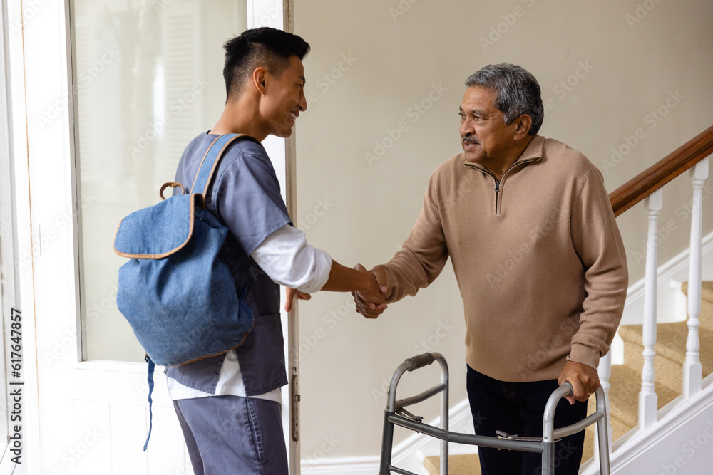 Fototapeta premium Diverse male doctor and senior male patient with crutches shaking hands at home