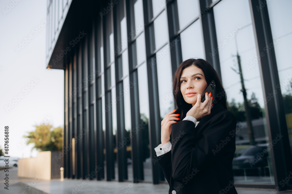 Happy young caucasian woman talking on phone standing near business center during day. Brunette wears strict clothes to work. Telephone technology concept Happy young caucasian woman talking on phone standing near business center during day. Brunette wears strict clothes to work. Telephone technology concept