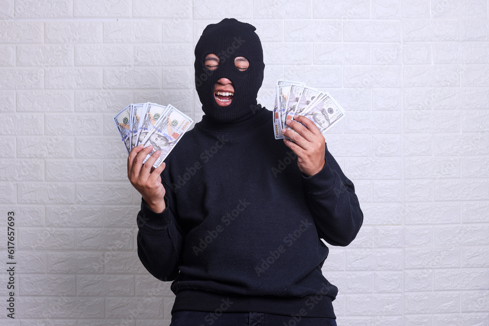 Robber man dressed in black hoodie stands with disguised face and holds a lot of money in his hands, stole a large amount, counts bills, isolated over gray background. Robber man dressed in black hoodie stands with disguised face and holds a lot of money in his hands, stole a large amount, counts bills, isolated over gray background.