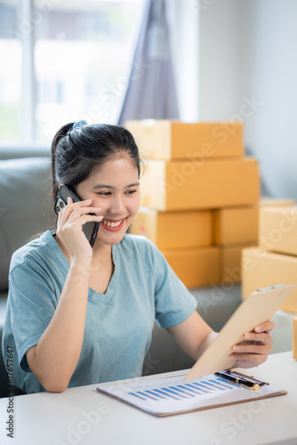 Happy young asian woman smiling at sales success after checking order from online store on smartphone at home office The concept of online commerce and e-commerce.