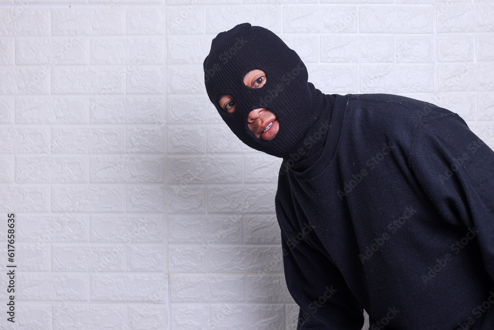 A criminal wearing black balaclavausing torch and peeking out from white wall A criminal wearing black balaclavausing torch and peeking out from white wall