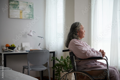 Sick senior woman sitting in wheelchair and looking through window