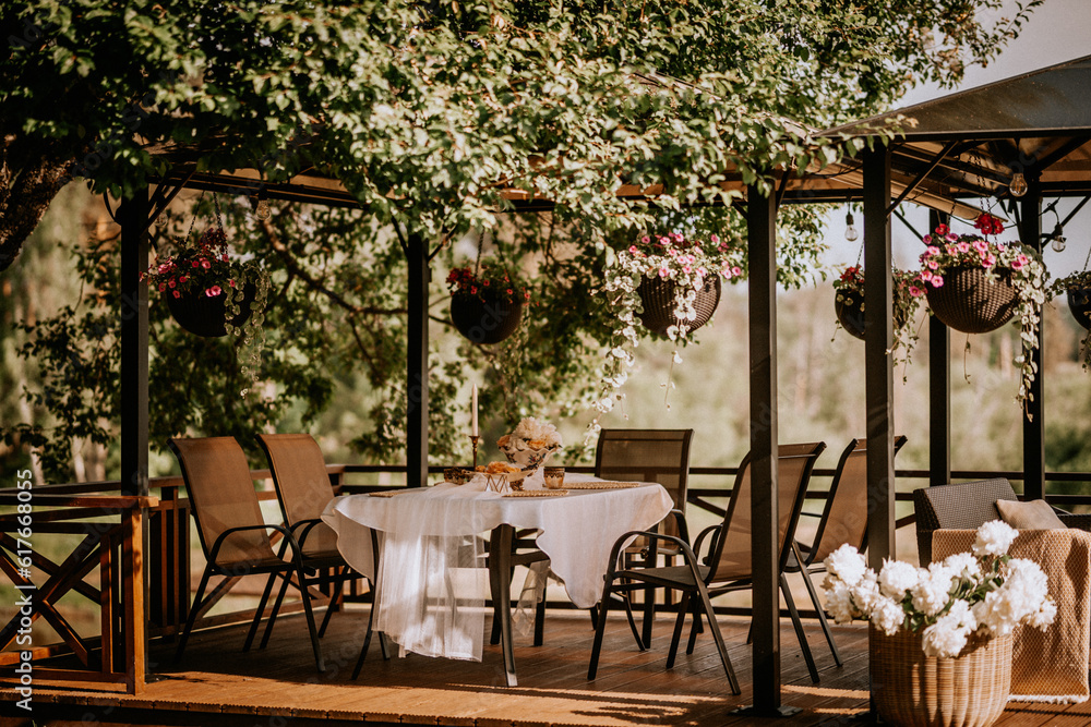 Fototapeta premium A white table is laid on the terrace and decorated with flowers in summer, which are hung in pots at sunset