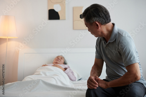 Senior man sitting on bed of sick wife sleeping in bed