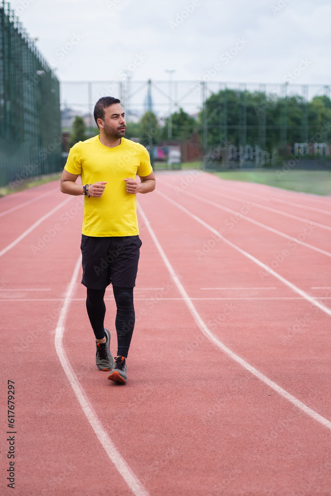 Man in tracksuit running along track with red rubber covering and white lines on outdoor sports arena in town park for health and body muscles development Man in tracksuit running along track with red rubber covering and white lines on outdoor sports arena in town park for health and body muscles development