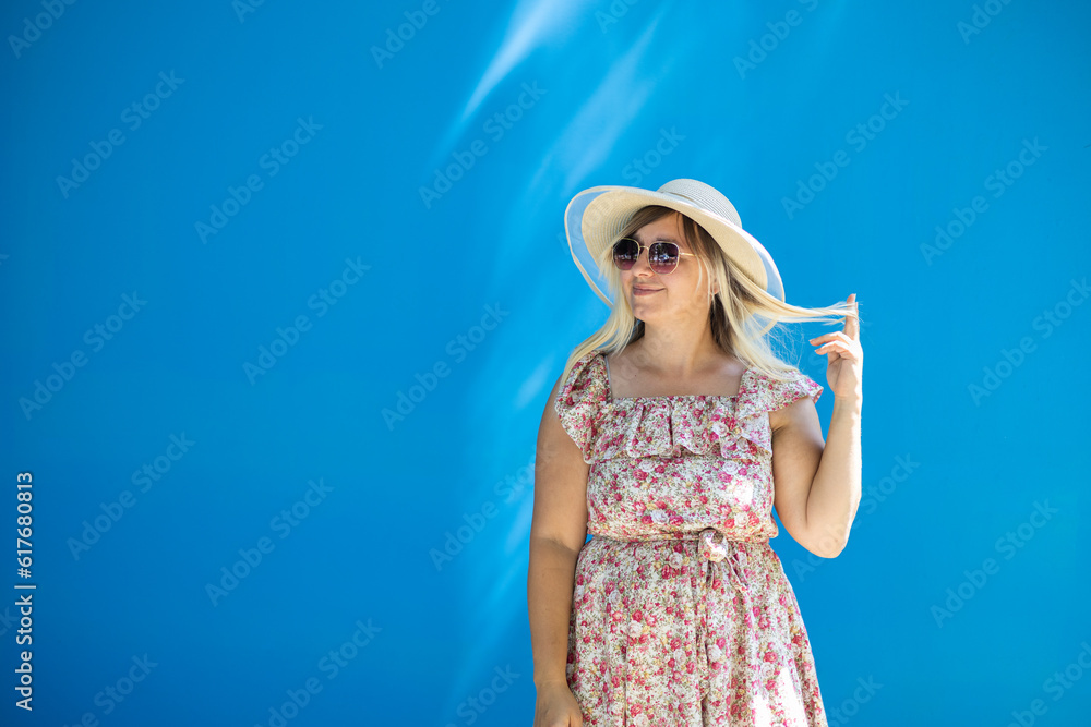 Beautiful woman in a hat, dress and sunglasses on a blue background in summer outdoors A woman twists a strand of hair around her finger and looks into the empty space on the side for inserting text. Beautiful woman in a hat, dress and sunglasses on a blue background in summer outdoors A woman twists a strand of hair around her finger and looks into the empty space on the side for inserting text.
