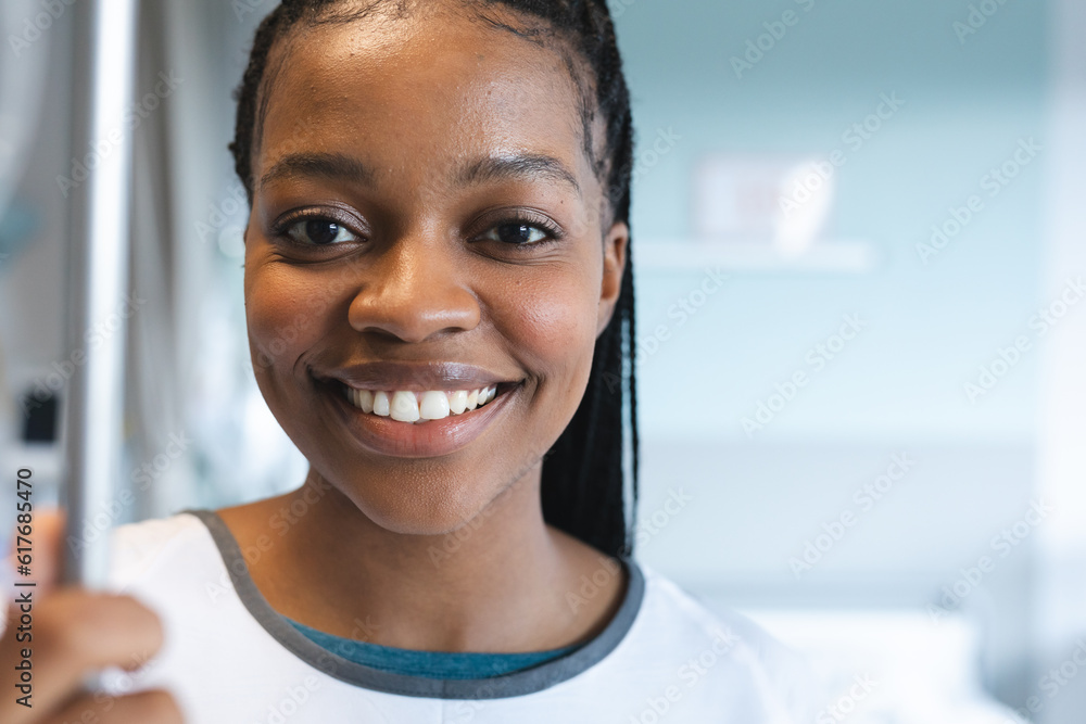 Portrait of happy african american female patient with braids, wearing hospital gown, holding drip