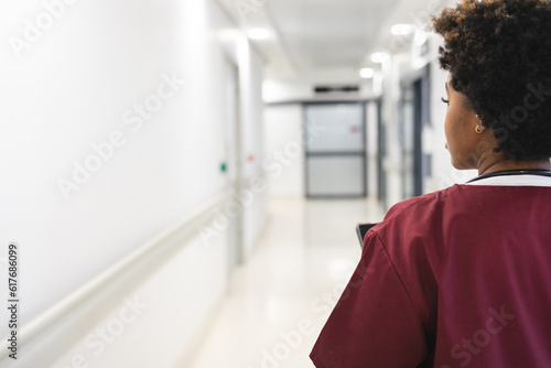 African american female doctor wearing scrubs, walking through corridor at hospital, copy space