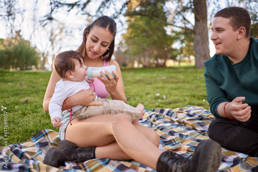 couple sitting on the lawn while the mother feeds her baby and her husband watches her couple sitting on the lawn while the mother feeds her baby and her husband watches her