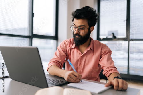 Portrait of bearded Indian businessman in glasses using laptop analyzing working project results, writing notes siting at table by window. Focused student male learning remotely looking to screen.