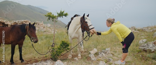 The woman feed domestic white horse on rocky steppe area on top of mountain. Girl looks and feeds horse with saddle green grass. Woman travels in summer, relaxing with nature. Landscape scenic view