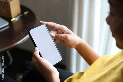 Closeup view of woman hands holding smartphone, sitting on bright living room. White screen for advertising text