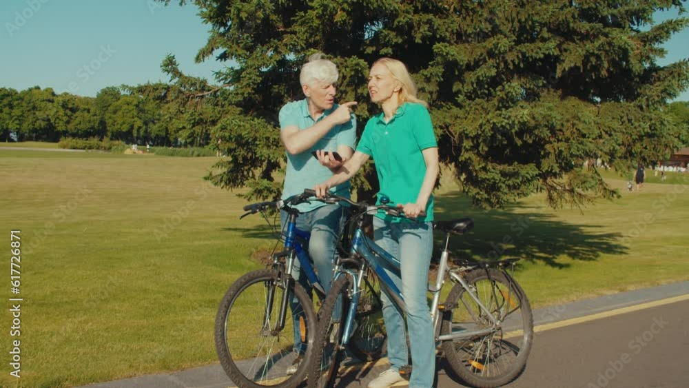Positive attractive senior couple of cyclists enjoying active lifestyle and summer vacations, discussing and choosing travel route using GPS navigation app on smartphone while riding bicycles in park Positive attractive senior couple of cyclists enjoying active lifestyle and summer vacations, discussing and choosing travel route using GPS navigation app on smartphone while riding bicycles in park