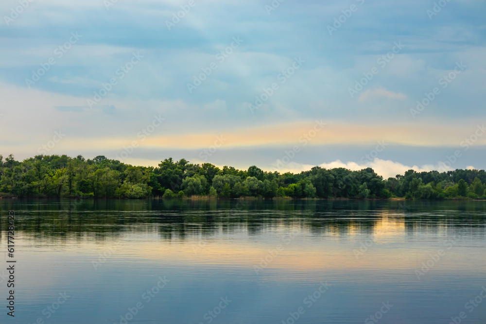 A picturesque landscape with a wide river, a forest on the shore and clouds in the sky on a summer day