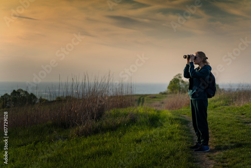Female hiker using binoculars for bird watching in green forest at sunset
