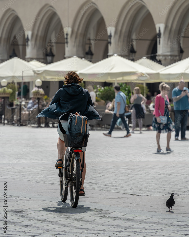 Authentic woman rides a bicycle in the old square on a sunny day against the background of tourists and umbrellas of a street cafe, a travel bag on the trunk of a bicycle, eco-friendly urban transport